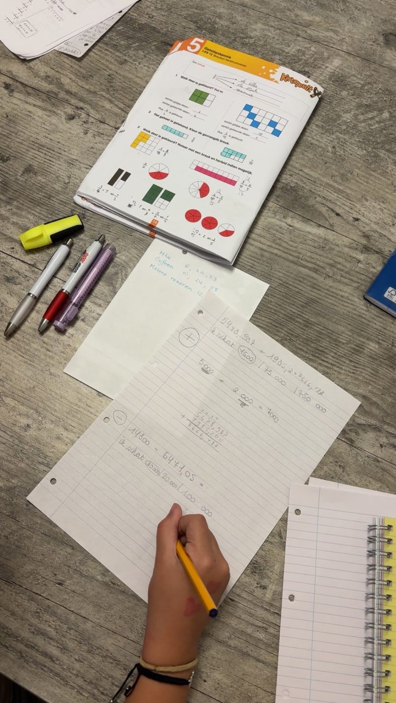 Hand van een kind dat met een geel potlood rekensommen op kladpapier maakt, met een open schoolboek erbij